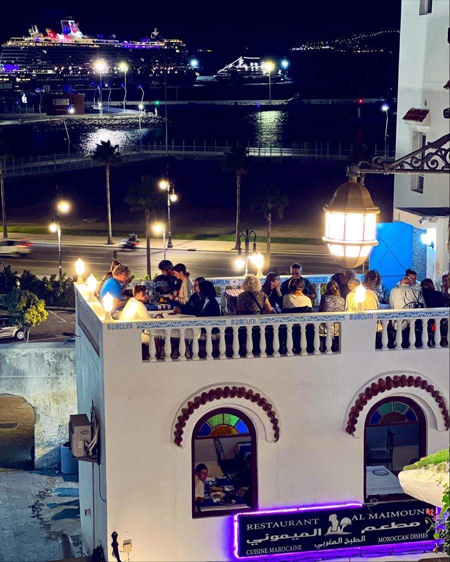 La terrasse un soir d'été — les convives sous la lanterne, le port et un paquebot illuminés derrière